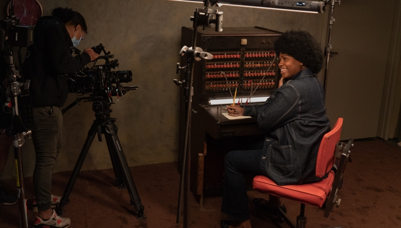A person with an Afro dressed in blue denim at a switchboard smiles at the camera while a film crew member checks the equipment