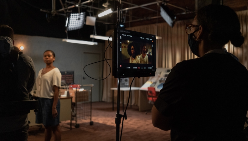 A masked crew member watches a monitor as several actors work on a scene