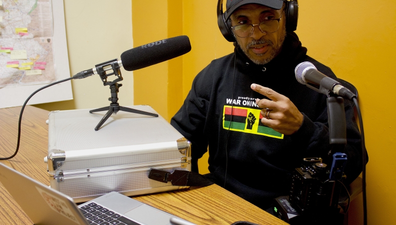 a man seated with two microphones pointing at him, making a point with one finger