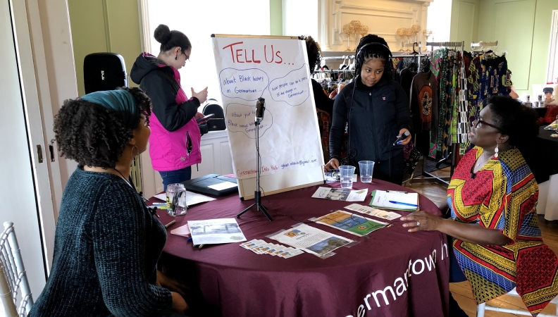 a woman seated at a table talking while a 2nd person holds a microphone and a 3rd person looks on
