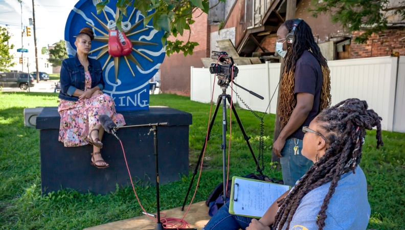 a woman sitting on a piece of public art while being interviewed by two people