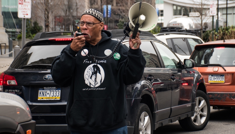A man wearing glasses, a black hoodie and a kufi hat stands amidst traffic while speaking into a bullhorn