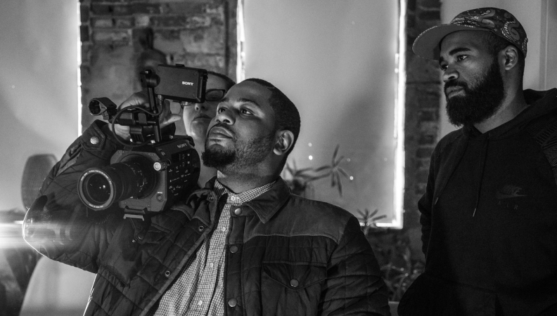 Three people looking at the viewfinder of a video camera one person has on their right shoulder