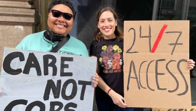 two people hold up signs that read "care not cops" and "24/7 access"