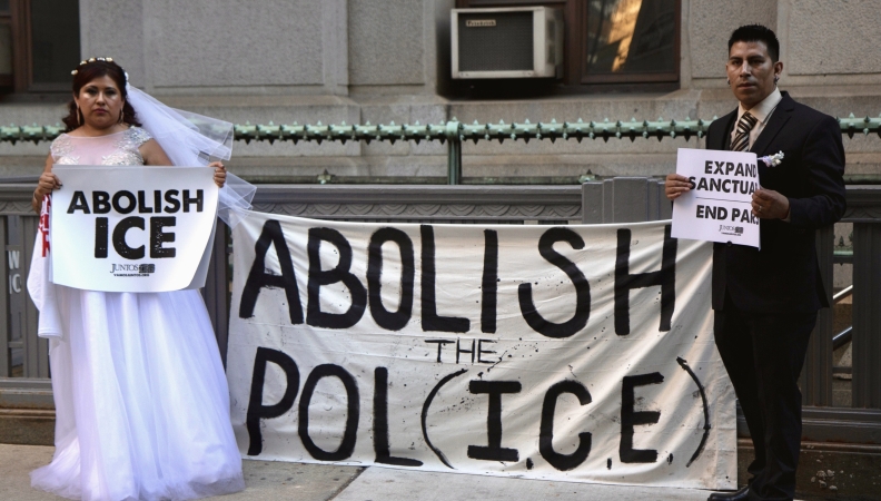 A bride and a groom each hold signs of protest against ICE while standing on opposite ends of a banner that reads "Abolish the Pol(ICE)"