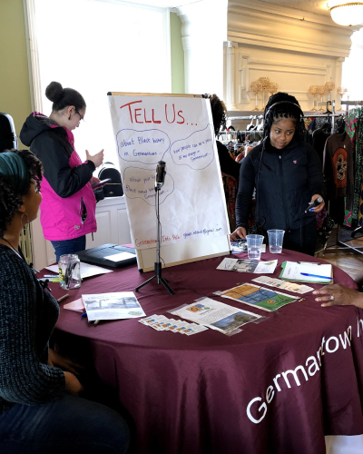 a woman seated at a table talking while a 2nd person holds a microphone and a 3rd person looks on