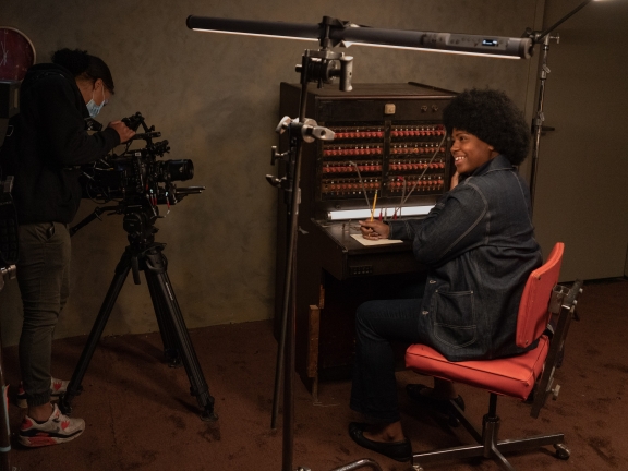 A person with an Afro dressed in blue denim at a switchboard smiles at the camera while a film crew member checks the equipment