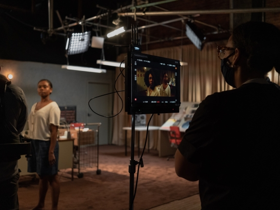 A masked crew member watches a monitor as several actors work on a scene