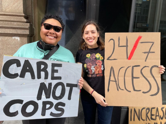 two people hold up signs that read "care not cops" and "24/7 access"