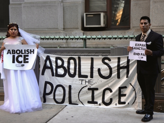 A bride and a groom each hold signs of protest against ICE while standing on opposite ends of a banner that reads "Abolish the Pol(ICE)"
