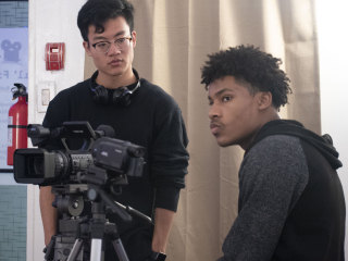 A light toned person wearing glasses and headphones looks at a sitting medium dark toned person with a video camera in the foreground