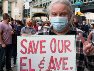 a man standing at a demonstration holding a sign saying "Save Our El & Ave" and flashing a peace sign