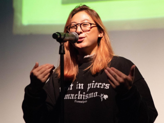 woman wearing glasses stands as she speaks into a microphone